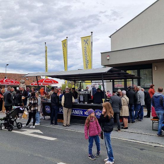 Eine Menschenmenge versammelt sich vor einem Imbissstand, mit Bannern und einem Schirm. Leute stehen Schlange, plaudern und einige tragen Jacken. Ein Kinderwagen und ein Kind sind im Vordergrund.