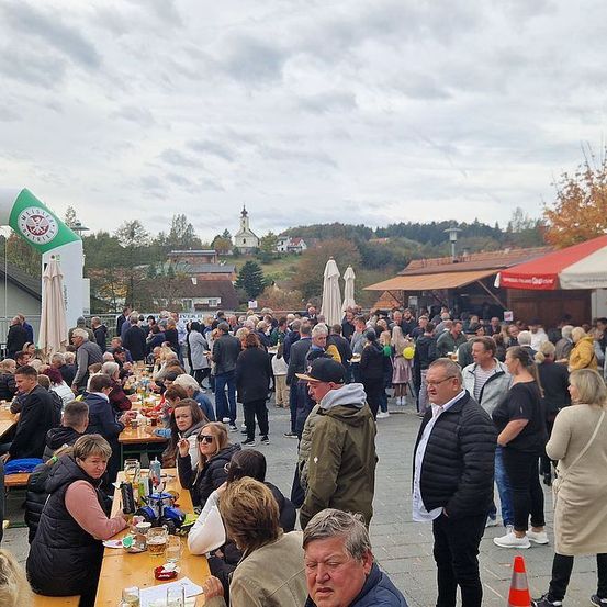 Eine große Menschenmenge speist im Freien bei einer Veranstaltung. Tische und Bänke sind mit Gästen gefüllt. Die Umgebung umfasst einen festlichen Torbogen und Essensstände. Der Himmel ist bewölkt.