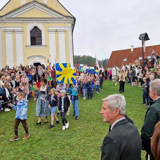 Eine große Menschenmenge, einschließlich Kinder, versammelt sich auf einem Rasenfeld, möglicherweise für eine Veranstaltung. Ein Gebäude mit einem Fenster und einer Flagge ist im Hintergrund.