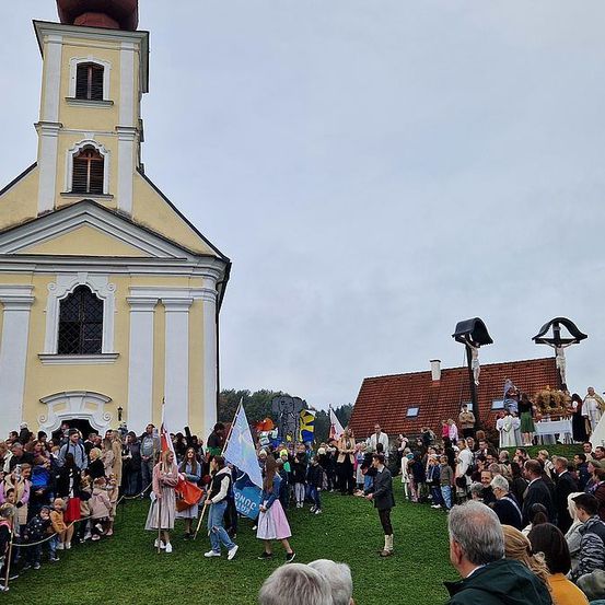 Eine große Menschenmenge versammelt sich vor einer Kirche. Einige Leute halten Fahnen. Ein Mann steht auf dem Rasen und hält eine Rede. Zwei Kreuze sind auf dem Dach eines Hauses platziert.
