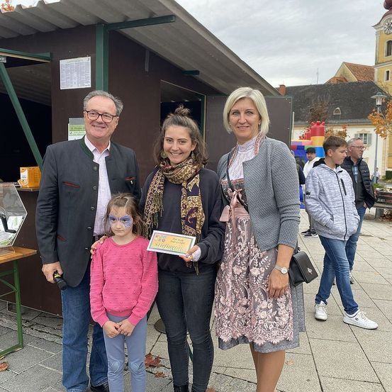 Eine vierköpfige Familie steht vor einem Gebäude und hält ein Zertifikat in der Hand. Das Gebäude hat ein Schild, und in der Nähe laufen zwei Jungen und ein Mann. Der Himmel ist bewölkt.