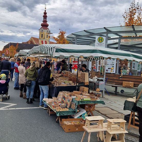 Ein geschäftiger Straßenmarkt mit Ständen und Menschen, die unter grün-weiß gestreiften Markisen stöbern. Eine Frau schiebt einen Kinderwagen, und eine Bank ist in der Nähe. Ein Turm und Bäume sind im Hintergrund.