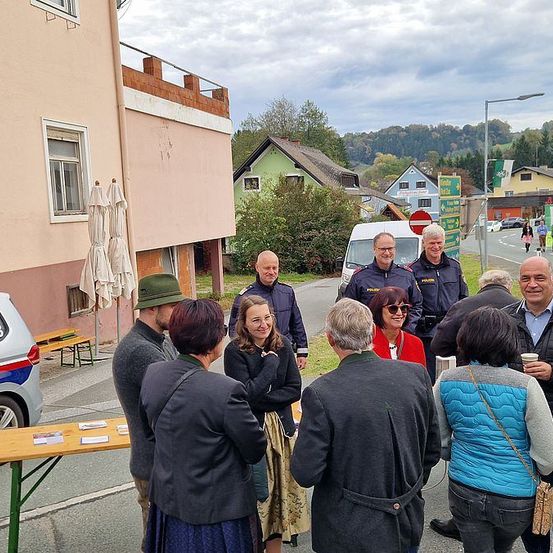 Eine Gruppe von Menschen in formeller Kleidung steht um einen Tisch an einer Straßenecke und unterhält sich. In der Nähe unterhalten sich zwei Polizisten in Uniform. Dahinter stehen Gebäude an der Straße, und in der Ferne fahren Autos vorbei.