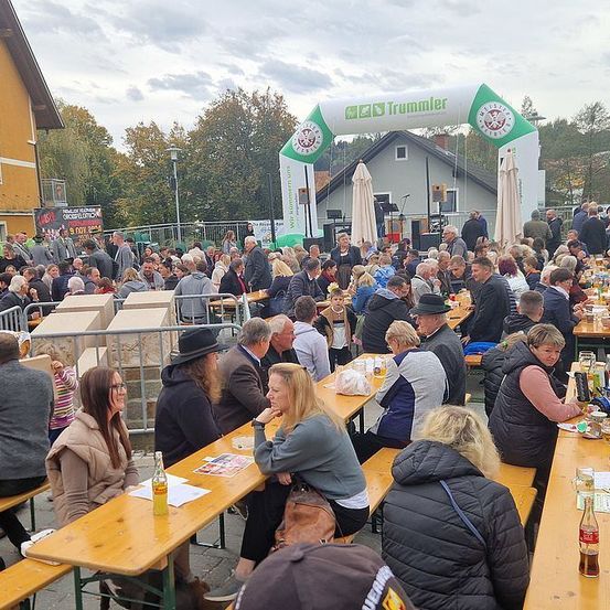Viele Menschen sitzen an Tischen im Freien. Sie befinden sich wahrscheinlich auf einem Festival, mit einem Bogen am Rücken mit dem Wort 'Trommeler'. Sie essen und trinken, einige mit Hüten.