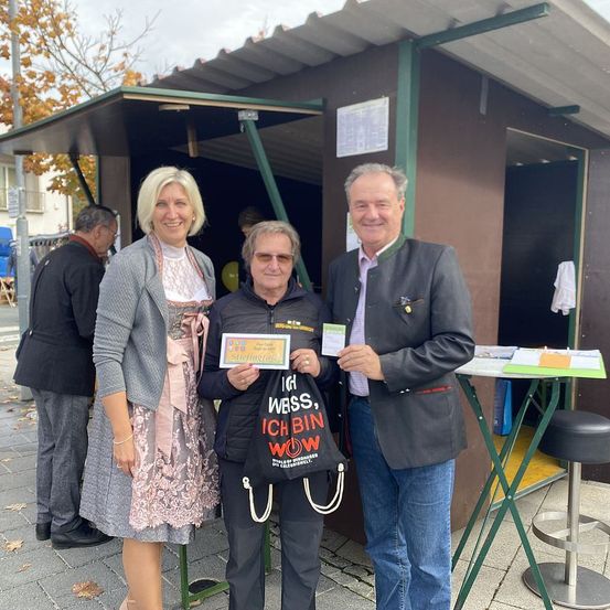 Drei Personen stehen vor einem Stand und lächeln. Der Mann rechts hält ein Schild und Papier, die mittlere Person einen Beutel, und die Frau links trägt ein Kleid. Hinter ihnen steht ein Mann bei einem Tisch mit einem Hocker.