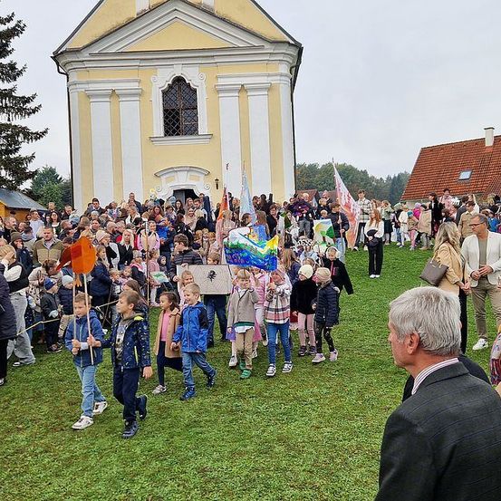 Eine Menschenmenge, darunter auch Kinder, versammelt sich vor einer Kirche und hält Flaggen und Schilder. Einige gehen, andere stehen. Ein Mann in Anzug schaut sie an.