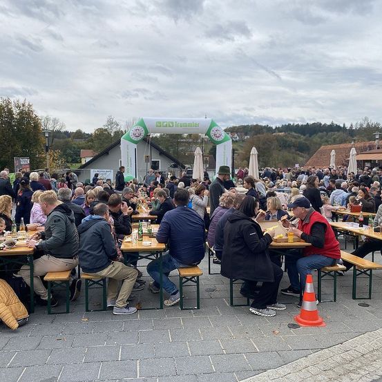 Eine große Menschenmenge sitzt an Tischen im Freien, einige essen. Der Bereich ist von Bäumen und Gebäuden umgeben, und es gibt ein großes Banner darüber.