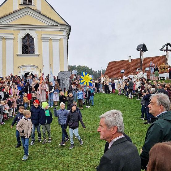 Eine große Menschenmenge versammelt sich vor einer Kirche für eine Veranstaltung, wobei Kinder laufen und Erwachsene stehen. Einige Personen tragen Mützen und tragen Fahnen. Dahinter ist ein großer Elefantenausschnitt zu sehen.