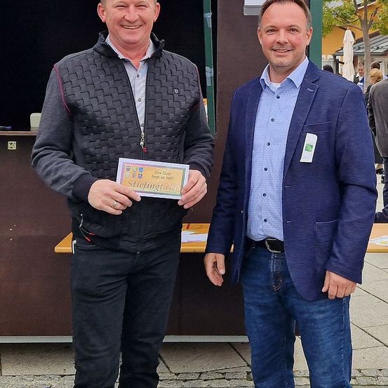 Zwei Männer stehen nebeneinander und lächeln für ein Foto. Der Mann auf der linken Seite hält ein gelbes Zertifikat mit einem Logo und Text. Hinter ihnen steht eine braune Wand mit einem Tisch.