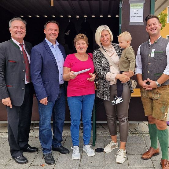 Fünf Menschen posieren für ein Foto im Freien. Ein Mann und eine Frau halten ein Papier. Eine Frau hält ein Baby. Sie alle lächeln. Dahinter steht ein Tisch mit einer grünen Wand.