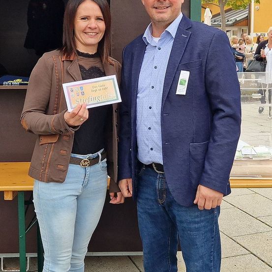 Eine lächelnde Frau in einer braunen Jacke hält ein Zertifikat in der Hand, während sie neben einem lächelnden Mann in einem blauen Blazer steht. Sie posieren für ein Foto im Freien.