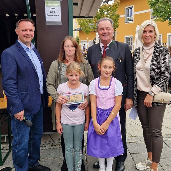 Fünf Erwachsene und zwei junge Mädchen stehen lächelnd im Freien, eine der Mädchen hält ein Zertifikat. Sie stehen vor einem gelben Gebäude, mit Bäumen und einem Tisch im Hintergrund.