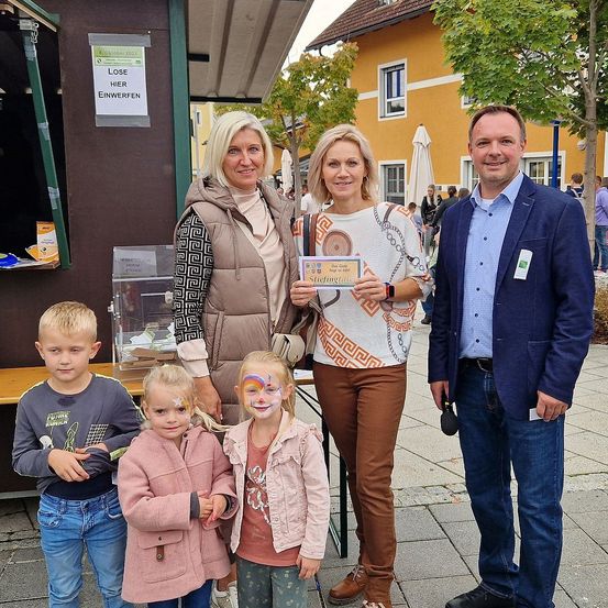 Vier Erwachsene und drei Kinder stehen gemeinsam auf einem Bürgersteig vor einem Gebäude. Sie lächeln und scheinen für ein Foto zu posieren. Hinter ihnen befindet sich ein Schild mit Text und ein kleiner Stand. Im Hintergrund sind ein Gebäude, Bäume und Menschen zu sehen, die auf der Straße gehen.