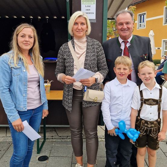 Vier Menschen stehen lächelnd zusammen draußen. Eine Frau hält ein Stück Papier in der Hand, während ein Mann ein traditionelles deutsches Outfit trägt. Zwei junge Jungen, einer mit einem Ballontier, stehen zwischen ihnen. Dahinter steht ein Tisch mit einem Becher und einem Schild mit deutschem Text.