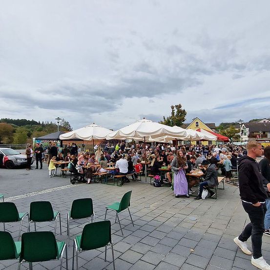 Ein großer Außenbereich mit vielen Menschen, die unter weißen Sonnenschirmen an Tischen sitzen. Einige Leute stehen und gehen. In der Ferne befinden sich Häuser und Autos.