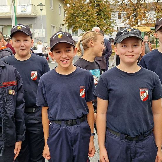 Eine Gruppe junger Menschen in marineblauen Uniformen, möglicherweise Feuerwehrleute, steht zusammen auf einem Außenbereich mit mehreren Personen dahinter und Gebäuden im Hintergrund.