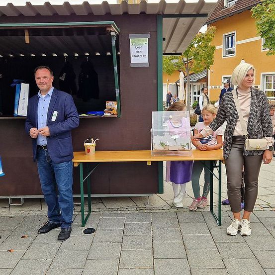Ein Mann und eine Frau stehen vor einem Stand mit einer Glasvitrine und einem Schild, begleitet von einem Kind. Sie befinden sich auf einem Stadtplatz, mit Gebäuden im Hintergrund.