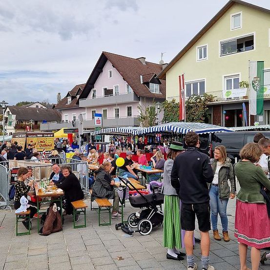 Ein geschäftiger Außenmarkt mit Menschen, die an Tischen unter gestreiften Zelten sitzen, einige mit Luftballons, vor Häusern.