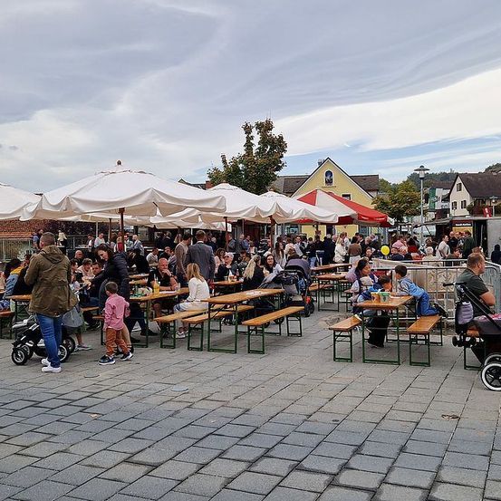 Ein Freiluft-Essbereich mit mehreren Tischen und Bänken, viele Menschen essen und trinken. Der Bereich ist mit weißen Sonnenschirmen bedeckt. In der Ferne befinden sich Häuser und ein bewölkter Himmel.