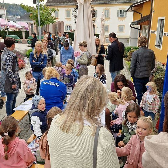 Eine Gruppe von Menschen, darunter Erwachsene und Kinder, versammelt sich in einer Stadt, einige mit Gesichtsbemalung. Eine Frau hält ein Baby, und ein Mann spricht mit einer Frau mit Brille. In der Nähe steht ein Kinderwagen.