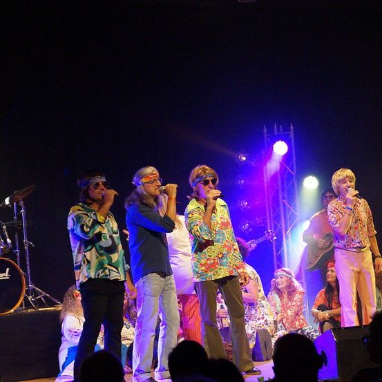 Eine Band tritt auf der Bühne mit farbenfrohen Outfits und Sonnenbrillen auf, umgeben von sitzenden Zuschauern und Bühnenbeleuchtung.