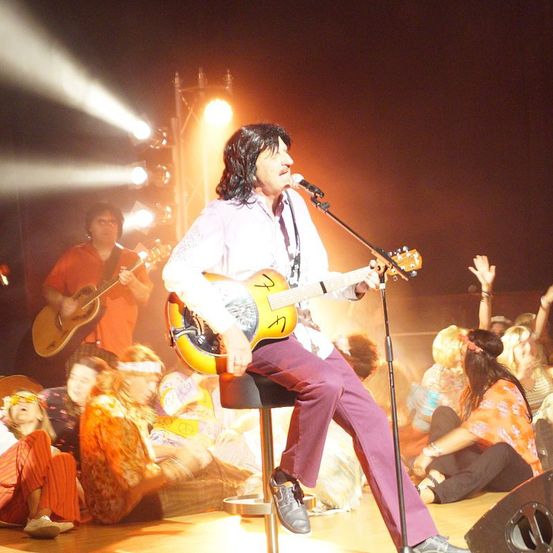 Ein Mann spielt auf der Bühne mit einer Gitarre und singt in ein Mikrofon, während er auf einem Hocker sitzt. Ein Bandkollege spielt Gitarre im Hintergrund. Das Publikum sitzt vor ihm auf dem Boden.
