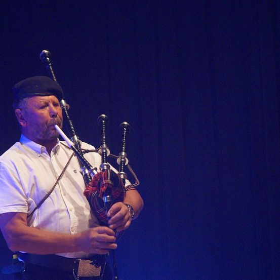 Ein Mann spielt auf der Bühne die Dudelsack, trägt ein weißes Hemd und eine schwarze Baskenmütze. Er hält das Dudelsack mit beiden Händen, und das Instrument hat eine rote gemusterte Abdeckung.