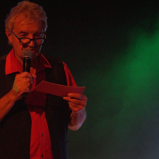 Ein älterer Mann mit Brille hält ein Papier und ein Mikrofon, wahrscheinlich bei einer Bühnenperformance. Er trägt ein rotes Hemd und eine schwarze Weste.