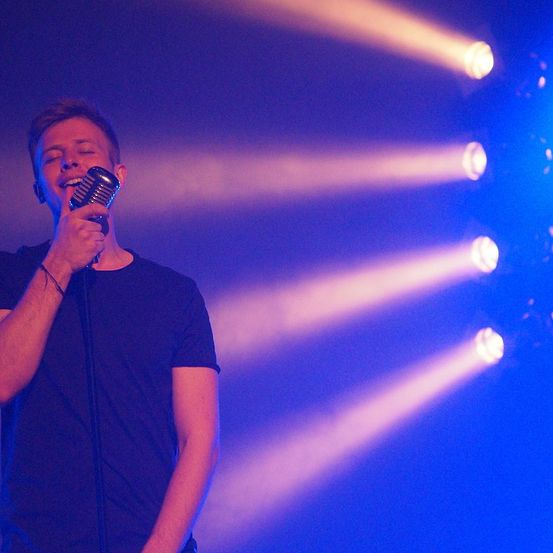 Ein Mann steht auf der Bühne und singt in ein Mikrofon mit geschlossenen Augen. Scheinwerfer sind auf ihn gerichtet, und er trägt ein blaues T-Shirt.