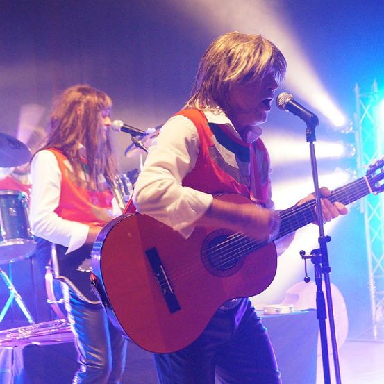 Zwei Musiker treten auf der Bühne auf; einer spielt Gitarre und singt, der andere spielt Trompete. Die Bühne ist mit blauen und violetten Lichtern beleuchtet.