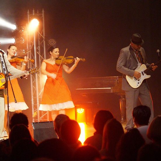 Eine Gruppe Musiker tritt auf einer Bühne mit Scheinwerfern auf. Die Geiger spielen, während der Gitarrist an der Seite steht. Das Publikum schaut von vorne zu.