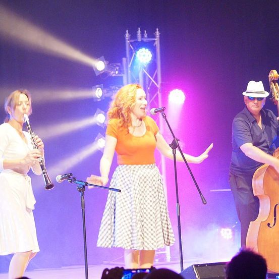 Ein Trio von Künstlern auf der Bühne, mit einer singenden Frau, einer Frau, die Klarinette spielt, und einem Mann, der Cello spielt. Die singende Frau trägt ein orangefarbenes Oberteil und einen karierten Rock. Die Bühne ist mit lila und weißen Lichtern beleuchtet.