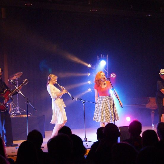 Eine Band spielt auf der Bühne, ein Mann spielt Gitarre und eine Frau Klarinette. Eine weitere Frau singt in ein Mikrofon. Die Bühne ist mit Scheinwerfern beleuchtet und hinter ihnen steht ein Klavier. Das Publikum beobachtet die Aufführung.