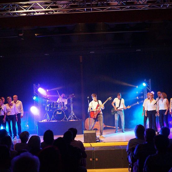 Eine Band tritt auf der Bühne auf, mit blauen und lila Beleuchtung. Der Leadsänger spielt Gitarre. Vor der Band stehen zwei Frauen und neben ihnen zwei Männer. Auf der linken Seite ist ein Schlagzeuger mit einem Schlagzeug-Set. Das Publikum sieht ihnen zu.