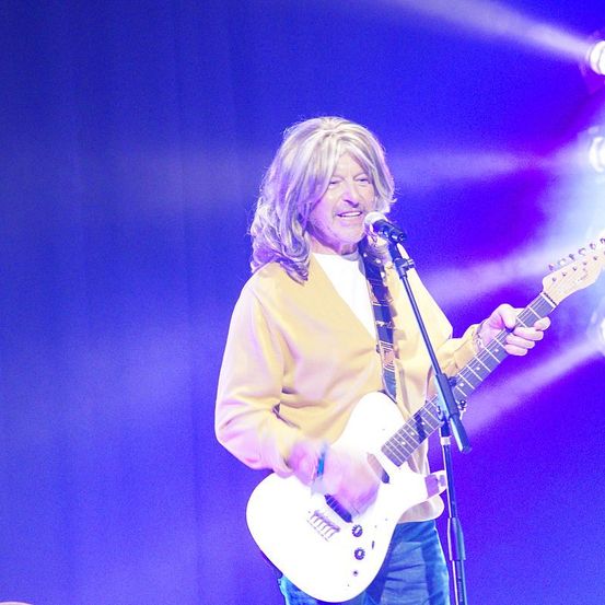 Ein Mann mit blonden Haaren spielt auf der Bühne eine weiße E-Gitarre. Er singt in ein Mikrofon.