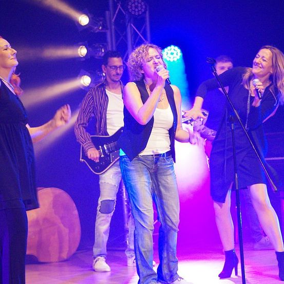Eine Band tritt auf einer Bühne mit farbiger Beleuchtung auf; zwei Frauen singen in Mikrofone, während ein Gitarrist hinter ihnen steht und eine Frau tanzt.
