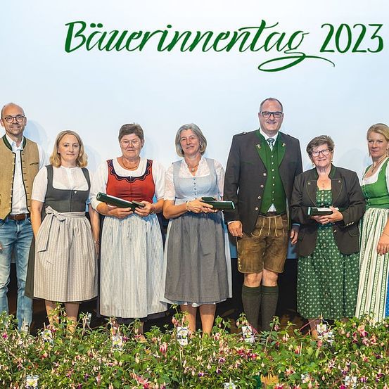 Eine Gruppe Erwachsener in traditioneller bayerischer Kleidung, die für ein Foto auf der Bühne mit Blumen vor einem weißen Hintergrund, der 'Bauerninnentag 2023' steht, posiert.