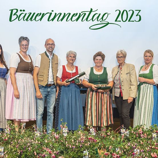 Eine Gruppe von Menschen steht in traditioneller bayerischer Kleidung, lächelt und zwei halten Auszeichnungen. Ein weißer Hintergrund trägt die Aufschrift 'Bauerninnentag 2023'.