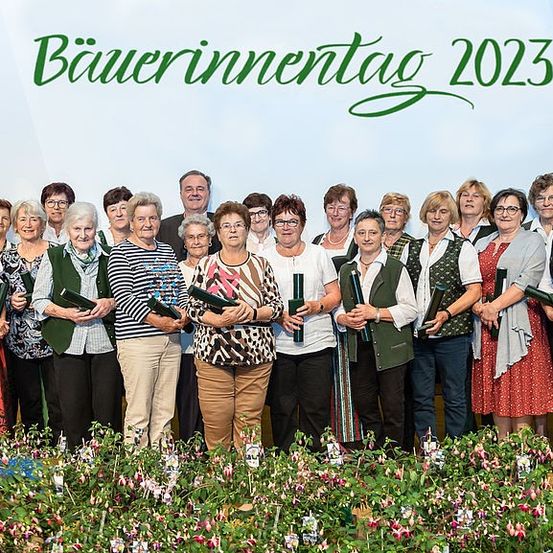 Eine Gruppe älterer Frauen und Männer in traditioneller bayerischer Kleidung posiert für ein Foto am Bayerischen Bauerntag 2023. Sie halten grüne Ordner in den Händen und stehen vor einem großen Schild.