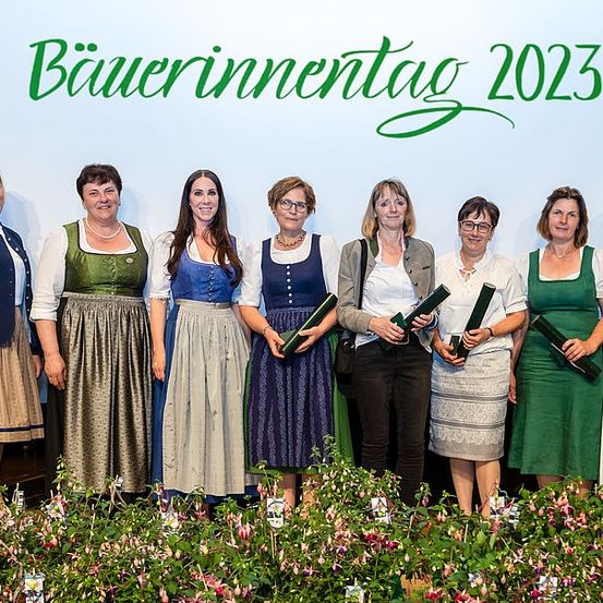 Eine Gruppe von Frauen in traditioneller Kleidung, die Urkunden halten, steht auf einer Bühne mit einem Blumenhintergrund und einem Banner mit der Aufschrift 'Bauerninnentag 2023'. Sie lächeln und posieren für ein Foto.