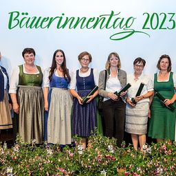 Eine Gruppe von Frauen in traditioneller Kleidung, die Urkunden halten, steht auf einer Bühne mit einem Blumenhintergrund und einem Banner mit der Aufschrift 'Bauerninnentag 2023'. Sie lächeln und posieren für ein Foto.
