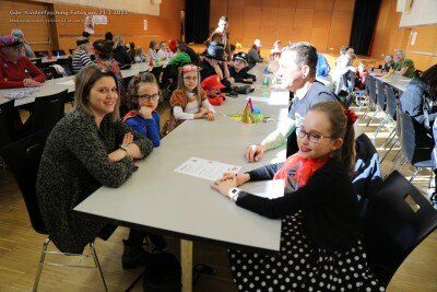 Eine Gruppe von Menschen, darunter Kinder in Kostümen, sitzen in einem Klassenzimmer an Tischen. Eine Frau und ein junges Mädchen schauen auf ein Blatt Papier.