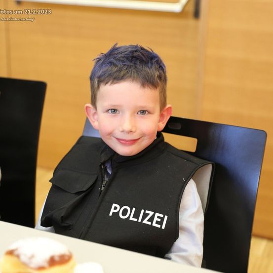 Ein Junge in einer schwarzen Polizeiweste mit weißem Text sitzt an einem Tisch mit einem Donut darauf.