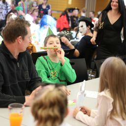 Eine Gruppe von Menschen auf einer Halloween-Party, ein Junge in Grün bläst eine lange Süßigkeit, ein Mann in Schwarz spricht mit ihm und ein Mädchen in Weiß hält einen Becher.