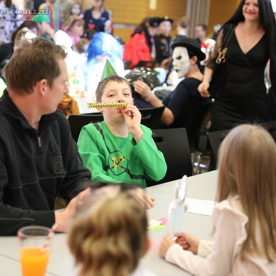 Eine Gruppe von Menschen auf einer Halloween-Party, ein Junge in Grün bläst eine lange Süßigkeit, ein Mann in Schwarz spricht mit ihm und ein Mädchen in Weiß hält einen Becher.