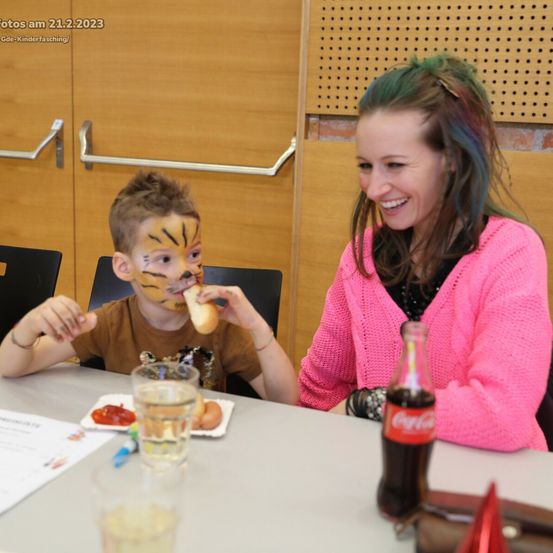 Ein Junge mit Tiger-Gesichtsbemalung isst Hot Dogs, während eine Frau ihn anlächelt. Ein Tisch hat Essen, Getränke und eine Coca-Cola-Flasche.