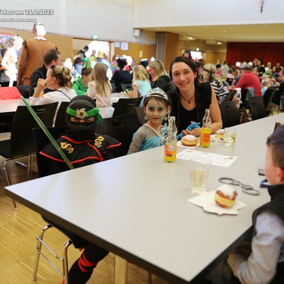 Eine Gruppe von Menschen sitzt an einem langen Tisch. Eine Frau lächelt und ein Kind ist in einem Ninja-Kostüm gekleidet. Der Tisch ist mit Essen, Getränken und verschiedenen Gegenständen gedeckt.