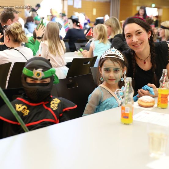 Eine Gruppe von Menschen, darunter eine Frau und zwei Kinder, sitzen an einem Tisch. Sie lächeln und posieren für ein Foto. Flaschen mit Getränken stehen auf dem Tisch. Dahinter sitzen auch andere Menschen.