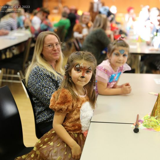 Ein Mädchen mit Gesichtsbemalung sitzt an einem Tisch mit anderen Kindern, eine erwachsene Frau beobachtet sie. Partyhüte und Dekorationen sind auf dem Tisch. Das Bild wurde am 21.2.2023 aufgenommen.