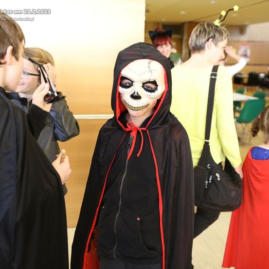 Ein junger Junge mit einer schwarzen Kapuze mit einem Totenkopfmaske steht unter anderen Leuten, einige in Kostümen, in einem Flur. Das Datum des Fotos ist 21.2.2023.